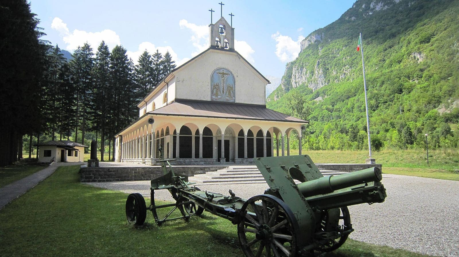 Il tempio ossario di Timau, frazione di Paluzza, con un cannone sul prato e le montagne intorno